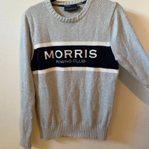 Grå stickad tröja Morris Rowing Club - Snygg grå stickad tröja från Morris Rowing Club med vit och marinblå rand över bröstet och broderad logga. Rund hals och ribbade muddar vid ärmslut och nederkant. Perfekt för dig som gillar klassisk stil med sportig touch.