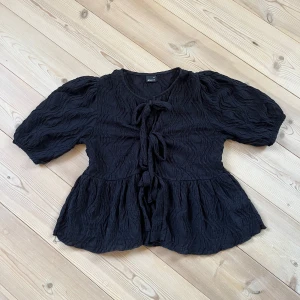 Svart blus med knyt från Gina Tricot - Supersöt svart blus från Gina Tricot i storlek XS. Den har puffiga korta ärmar, coolt strukturerat tyg och flera knyt framtill. Blusen har en lätt volang nertill och känns både trendig och unik. Perfekt att styla med jeans eller kjol för en snygg look.