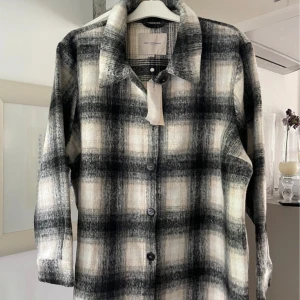 Rutig overshirt från Only Carmakoma - Overshirt i svart, grått och beige rutigt mönster från Only Carmakoma. Jackan har klassisk krage, knäppning framtill med svarta knappar och lång ärm. Materialet är mjukt och ulligt, perfekt för lager-på-lager. Snygg till jeans eller leggings.