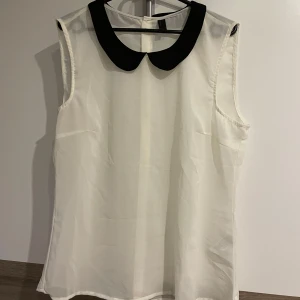 Vit blus med svart krage - Säljer en söt vit blus med ärmlös design och en kontrasterande svart, rundad krage. Blusen har en lätt och luftig känsla, perfekt för att skapa en stilren look. 