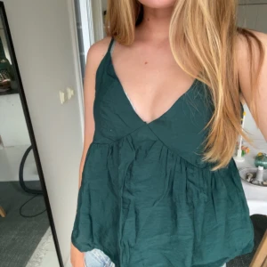 Mörkgrön omlott linnetopp från Aware - Supersöt mörkgrön linnetopp från Aware med omlott framtill och justerbara smala axelband. Toppen har v-ringning och lätt rynkad, luftig passform som ger en avslappnad vibe. Perfekt till jeansshorts eller kjol för en chill look.Den har en dragkedja på sidan av. 
