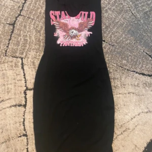 Svart kortklänning med tryck från SHEIN - Cool svart kortklänning från SHEIN i storlek XS. Framsidan har ett stort tryck med texten 'STAY WILD FANTASIA' och en örn i brunt, vitt och rosa. Klänningen är ärmlös och har rund hals. Perfekt för dig som gillar edgy streetstyle.