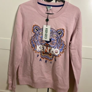 Rosa Kenzo sweatshirt med tiger - Säljer en rosa sweatshirt från Kenzo i storlek S med det ikoniska broderade tigermotivet i blått, orange och vitt på bröstet. Tröjan har rund hals, långa ärmar och ribbade muddar. Perfekt för dig som gillar statement-plagg och coola designer.