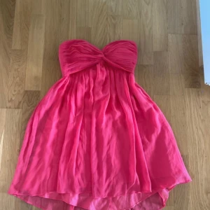 Axelbandslös rosa kortklänning - Säljer en axelbandslös kortklänning i stark rosa färg med draperad överdel och lätt volang i kjolen. Klänningen har en snygg omlott-effekt upptill och är perfekt för sommarens alla tillfällen. Materialet känns lätt och luftigt.