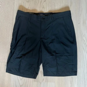 Svarta shorts från Greg Norman - Snygga svarta shorts från Greg Norman med stretchig midja och klassisk passform. De har framfickor, bakfickor med knapp och bälteshällor. Materialet är mjukt och lätt polyester, perfekt för en chill sommardag. Diskret logga på bakfickan och stilren design.