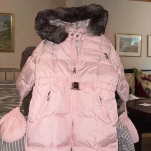 Ljusrosa dunjacka barn med päls från Armani - Storlek 62! Säljer en ljusrosa dunjacka från Armani med fluffig mörk päls på huvan, snyggt bälte med metallspänne och flera dragkedjor. Jackan har praktiska fickor och medföljande matchande vantar. 