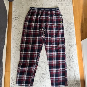 Lyle & Scott PyjamasByxor - Mysiga rutig pyjamasbyxor från Lyle & Scott med elastisk midja och logotyp. Byxorna har vinrött, svart och vitt rutmönster samt en liten gul broderad logga. Tillverkade i mjuk bomull, perfekta för chill hemma.