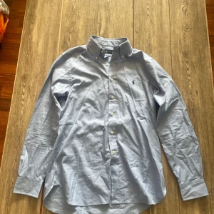 Blå skjorta från Ralph Lauren - Snygg blå skjorta från Ralph Lauren med klassisk krage och knappar framtill. Skjortan har ett diskret rutmönster och den ikoniska broderade loggan på bröstet. Långärmad och tillverkad i mjuk bomull som känns skön mot huden. Storlek L men känns mera som XL