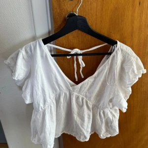 Vit volangtopp med knytband - Säljer en söt vit topp med volanger och v-ringning. Toppen har korta ärmar och knytband i ryggen för extra detalj. Perfekt för varma dagar och enkel att matcha med jeans eller kjol.