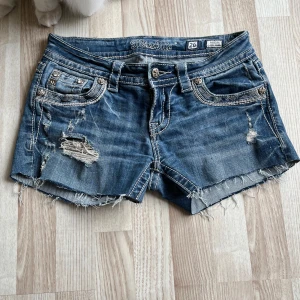 Miss me jeansshorts med broderade bakfickor - Snygga blå jeansshorts från Miss Me med slitna detaljer. Bakfickorna har broderade mönster med inslag av blommigt tyg och paljetter. Klassisk femficksmodell med låg midja och dekorativa sömmar. Helt utan defekter i storlek 26                                                            Midjemått: 36 cm Grenens längd: 18 cm Hela shortsens längd: 25 cm