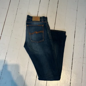 Blå jeans från Nudie Jeans - Säljer ett par klassiska blå jeans från Nudie Jeans med orangea sömmar och fem fickor. Jeansen har normal passform och är tillverkade i denim med snygg tvätt och slitningar. Perfekta för en avslappnad stil. Storlek W30 L 32