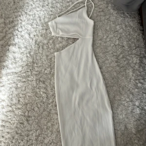 Vit ribbad långklänning med oneshoulder - Säljer en vit ribbad långklänning med smala axelband och oneshoulder-design. Klänningen har en snygg cut-out vid midjan och är figurnära. Perfekt för dig som vill sticka ut med en modern och trendig look.