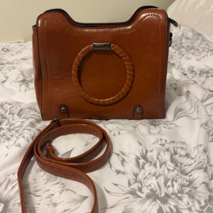 Brun handväska med rund handtag - Säljer en snygg brun handväska i skinnimitation med ett unikt runt handtag och avtagbar axelrem. Väskan har dragkedja upptill och en mindre ficka med dragkedja på baksidan. Perfekt för dig som vill ha en stilren accessoar.