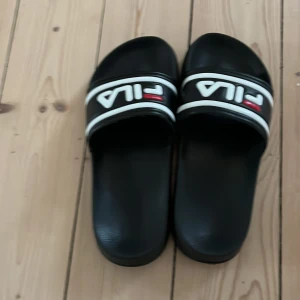Svarta sandaler från Fila - Svarta sandaler från Fila med vit och röd logga på remmen. Klassisk slip-on modell med bred rem över foten och platt sula. Perfekta för sommaren eller att ha hemma.