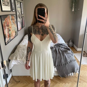 Vit klänning med fina detaljer på ryggen  - En vit klänning med smala axelband och v-ringning. Klänningen har en figurnära passform upptill och utställd kjol, samt korsade band i ryggen. Perfekt för studentklänning eller till midsommar. Se bild på fläckar på INSIDAN av bysten, syns alltså inte vid användning 