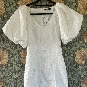 Vit kortklänning med puffärmar  - Superfin vit kortklänning från Gina Tricot med stora puffärmar och v-ringning. Klänningen har en figurnära passform och är tillverkad i ett strukturerat, lätt glansigt material som ger en lyxig känsla. Perfekt för sommarens speciella tillfällen som studenten! Helt ny, köpte den när dom släpptes men känner att den är lite liten för mig och kan inte längre lämna tillbaka.. 