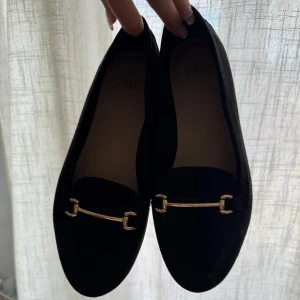 Svarta loafers med gulddetalj - Snygga svarta loafers med guldfärgad detalj framtill. Perfekta för en stilren look och passar till många olika outfits. Skorna har en klassisk design med en modern touch. 