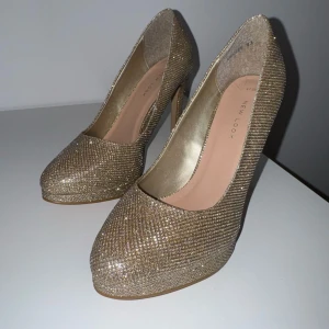 Glittriga guldiga pumps från New Look - Säljer ett par glittriga pumps i guld från New Look. Skorna har rund tå, hög klack och är täckta av glittrigt material som verkligen sticker ut. 