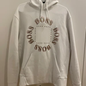 Vit hoodie från Hugo Boss - Säljer en vit hoodie från Hugo Boss med stor BOSS-logga i brunt och beige framtill. Tröjan har huva med dragsko och en stor magficka. Perfekt för en avslappnad och stilren look.