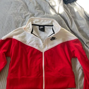 Träningsjacka från Nike - Säljer en knappt använd, snygg röd och vit jacka från Nike med dragkedja framtill och hög krage. Jackan har svart Nike-logga på bröstet och är perfekt för en sportig stil. Passar dig som gillar streetwear och vill ha något bekvämt och stilrent. Lite kortare modell. Kan mötas i Gbg :)