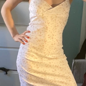 Vit blommig omlottklänning - Söt vit klänning med små blommor i rosa och grönt. Klänningen har omlott-skärning upptill och är ärmlös med smala axelband. Passformen är tight och tyget är mjukt och lätt, perfekt för varma dagar.