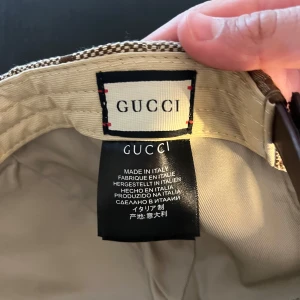 Beige och brun keps från Gucci - Snygg keps från Gucci i beige och brunt med stort GG-mönster och justerbar rem baktill i brunt läder. Klassisk böjd skärm och exklusiv känsla, perfekt för dig som gillar designeraccessoarer.