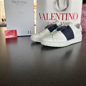 Valentino Garavani Open sneakers med blå detalj - Säljer ett par vita Valentino Garavani Open sneakers med bred blå rem över mitten. Skorna har klassisk rund tå, platt sula och vita snören. Kommer med originalkartong och påse. Perfekta för dig som gillar stilrena och exklusiva sneakers. Storlek 44 men passar på 43 ända skadan på boxen är lappen som ni ser på bilden