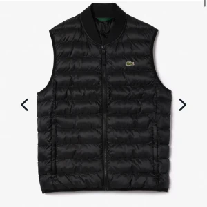 Svart väst från Lacoste - Snygg svart dunväst från Lacoste med dragkedja framtill och klassisk logga på bröstet. Västen är lätt och har en quiltad design, perfekt för lager-på-lager. Fodrad med Thermore för extra värme.