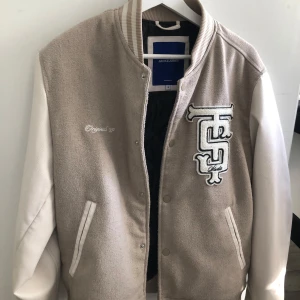 Beige och vit collegejacka från Jack & Jones - Säljer en snygg beige och vit collegejacka från Jack & Jones med broderad logga på bröstet och texten 'Original 90'. Jackan har långa vita ärmar, ribbade muddar och knäppning framtill. Finns även en innerficka vilket som är extra bra att förvara saker i. Storleken är S. Original pris är 1000 kr