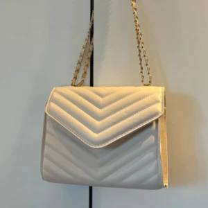 Beige  axelväska med kedjerem - Säljer en stilren beige axelväska med quiltat mönster och guldfärgad kedjerem. Väskan har ett lock med magnetlås och insidan är svart. Perfekt för att lyfta din outfit med en elegant touch.