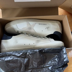 Golden Goose Superstar sneakers i vitt och grått - Golden Goose Superstar sneakers i vitt skinn med grå mockadetaljer och stjärna på sidan. Skon har grå snörning, svart hälparti och en platt sula. Klassisk och stilren modell med diskreta logodetaljer. Används en gång