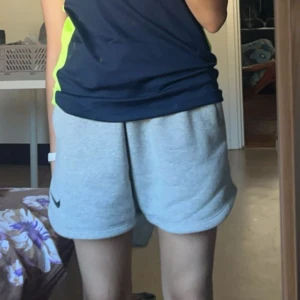 Grå sweatshorts från Nike - Säljer ett par grå sweatshorts från Nike med svart snörning i midjan och klassisk Nike-logga på benet. Shortsen har en loose fit och är tillverkade i mjuk bomullsmix, perfekta för träning eller chill. Har två fickor med dragkedja 