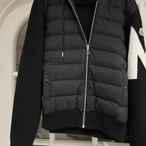 Moncler jacka | as fet | ev byte - Säljer en svart pufferjacka från Moncler med huva och dragkedja. Jackan har quiltad framsida, stickade ärmar och stor vit Moncler-logga på ena ärmen. Snygg och sportig design med fickor framtill. | EV BYTE 