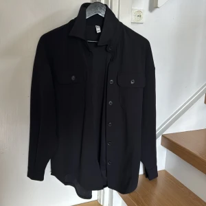 Svart overshirt från JDY - Säljer en svart skjorta från JDY med klassisk krage och två bröstfickor med knappar. Skjortan har långa ärmar och knäppning framtill. Perfekt för en stilren look.