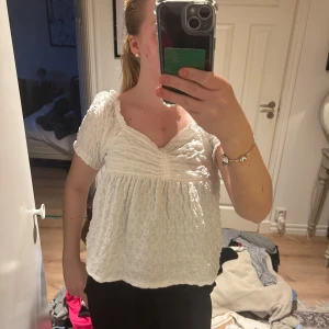 Vit blus från Gina Tricot - Supersöt vit blus från Gina Tricot med korta puffärmar och smockad struktur. Djup v-ringning och lätt volang vid ärmslut och nederkant. Perfekt till sommaren och enkel att styla med jeans eller kjol.