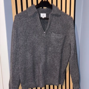 Grå stickad tröja med dragkedja från H&M - Mysig grå stickad tröja från H&M med halv dragkedja framtill och krage. Tröjan har lång ärm och en liten bröstficka. Perfekt för lager-på-lager och enkel att matcha med det mesta.