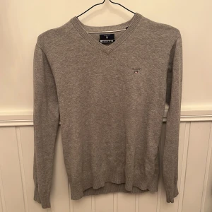 Grå v-ringad tröja från GANT - Säljer en klassisk grå v-ringad tröja från GANT i bomull. Tröjan har lång ärm, ribbade muddar och en liten broderad GANT-logga på bröstet. Perfekt för en stilren och enkel look.