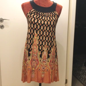 Mönstrad ärmlös klänning från Fashion In Style - Säljer en ärmlös klänning från Fashion In Style med färgstarkt mönster i svart, orange och gult. Klänningen har öppen rygg med knytband i nacken och är tillverkad i viskosblandning. Perfekt för dig som gillar unika prints och bekväma material.