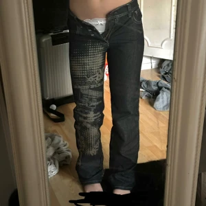 Mörkblå jeans med grafiskt tryck - Säljer ett par mörkblå jeans med unikt grafiskt tryck i beige på vänster ben och bakficka. Jeansen har normal passform och raka ben. Snygga kontrastsömmar och klassisk femficksmodell.