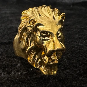 Kraftfull lejonring i guldfärgat rostfritt stål – retrodesign med attityd - Uttrycksfull ring i guldfärgat rostfritt stål med detaljerat lejonhuvud i 80-talsstil – en riktig poweraccessoar. Innerdiameter ca 2 cm, höjd ca 3,5 cm. Mycket lite använd, i utmärkt skick. Ingen prutning – bundlerabatt erbjuds vid köp av flera varor! 