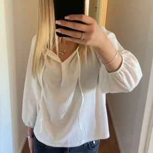 Vit blus med trekvartsärm - Säljer en stilren vit blus med trekvartsärmar och v-ringning. Blusen har knytband vid halsen och är tillverkad i ett lätt, luftigt material. Perfekt för en fräsch och enkel look.