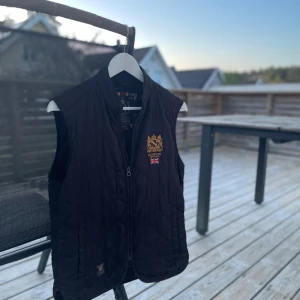 Svart quiltad väst från Morris - Säljer en svart quiltad väst från Morris med ståkrage och dragkedja framtill. Västen har broderad logga i guld på bröstet och klassisk brittisk stil. Perfekt för lager-på-lager och snygg till många outfits. Hål i ena fickan och mini slitage på ryggen