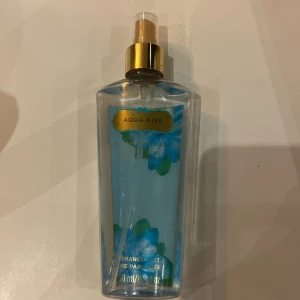 Aqua Kiss body mist från Victoria's Secret - Fräsch body mist med blå vätska och blommigt motiv på flaskan. Flaskan är genomskinlig med gulddetaljer på spraymunstycket och har en elegant, modern design. Doften är lätt och uppfriskande, perfekt för dig som gillar fräscha toner. Köpt för länge sedan