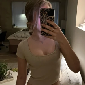 Beige ribbad topp från Lindex - Säljer en beige ribbad topp från Lindex med korta ärmar och rundad, djup urringning. Toppen är i mjuk bomull och har en tight passform som framhäver figuren. Perfekt att matcha med jeans eller kjol.