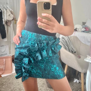 Turkos glittrig minikjol med volang - Helt oanvänd, ny med prislapp! Säljer en turkos glittrig minikjol med stora volangdetaljer framtill. Kjolen har en strukturerad yta och en tight passform som framhäver siluetten. Perfekt för dig som vill sticka ut med en färgstark och unik look.