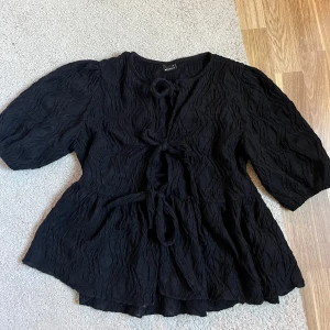Svart blus med knyt från Gina Tricot - Säljer en svart blus från Gina Tricot i storlek XS. Blusen har tre knytningar framtill, puffiga trekvartsärmar och ett vågigt, strukturerat tyg. Perfekt för dig som gillar trendiga detaljer. Inte använd alls mycket. 