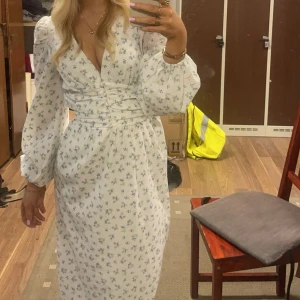 Vit blommig omlottklänning med puffärm - Söt vit klänning med små blå blommor, omlottliv och markerad midja. Långa puffärmar och v-ringad hals ger en romantisk känsla. Perfekt för vår och sommar!