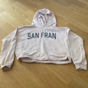 Ljusrosa hoodie med SAN FRAN-tryck från H&M - Säljer en ljusrosa hoodie från H&M med trycket 'SAN FRAN' i svart på bröstet. Tröjan har huva och långa ärmar, samt en croppad passform som ger en trendig look. Perfekt för en avslappnad stil.