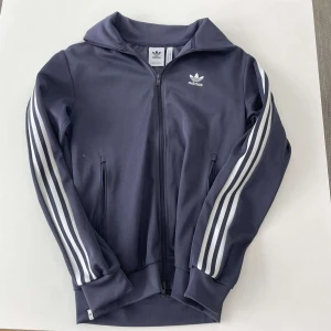 Adidas firebrid, mörkblå Adidas track jacket med vita ränder - Säljer en klassisk mörkblå Adidas track jacket med vita ränder längs ärmarna och dragkedja framtill. Jackan har ståkrage, två fickor med dragkedja och Adidas logga på bröstet. Perfekt för en sportig och avslappnad stil. Väldigt bra skick, pris kan diskuteras vid snabb affär!
