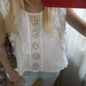 Vit blus med broderade detaljer - Säljer en söt vit blus med broderade blommönster framtill och volangdetaljer längs axlarna. Blusen är ärmlös och har en luftig passform, perfekt för varma dagar.